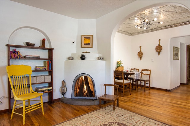 A cozy living area features a traditional kiva fireplace with a warm glow. Curved archways lead to a dining space, adorned with wooden chairs and a light fixture radiating elegance. A bookshelf, complemented by a bright yellow chair, adds a touch of color to the wood-floored space.