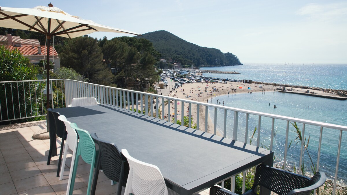 A spacious terrace is furnished with a large table and colorful chairs, overlooking a beach with gentle waves. The view includes hills in the background and a serene bay, inviting relaxation and outdoor dining.