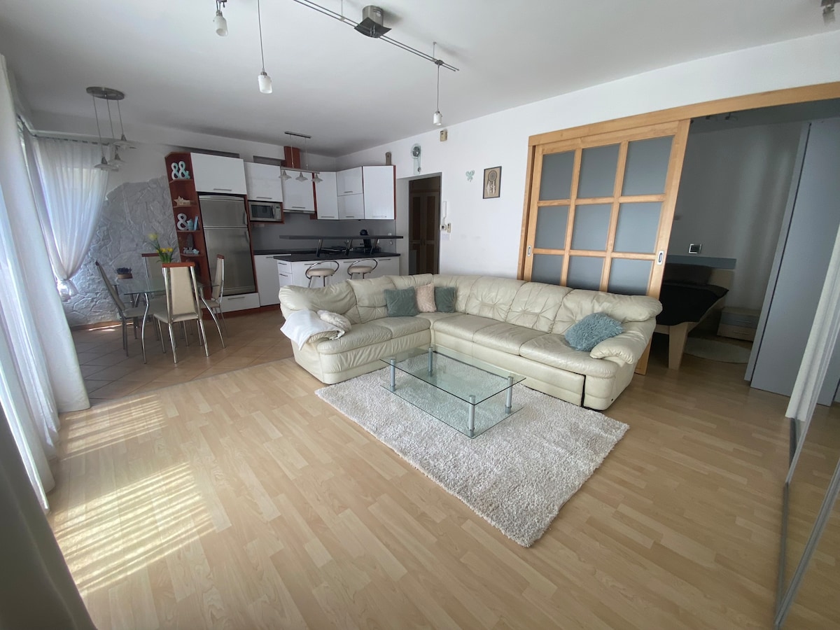 A spacious living area features a white sectional couch and a glass coffee table, complemented by a light-colored rug. Natural light enters through large windows, enhancing the open layout that connects to a well-equipped kitchen in the background.