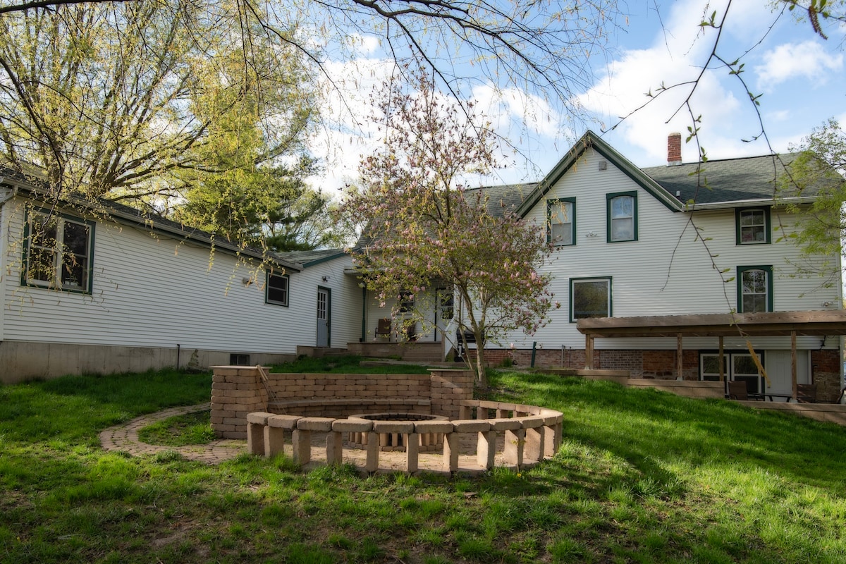 A spacious backyard is featured, highlighted by a circular stone fire pit surrounded by seating. A well-maintained lawn provides ample green space, while a blooming tree adds a touch of nature. The exterior of the house is visible, showcasing multiple windows and a porch area.