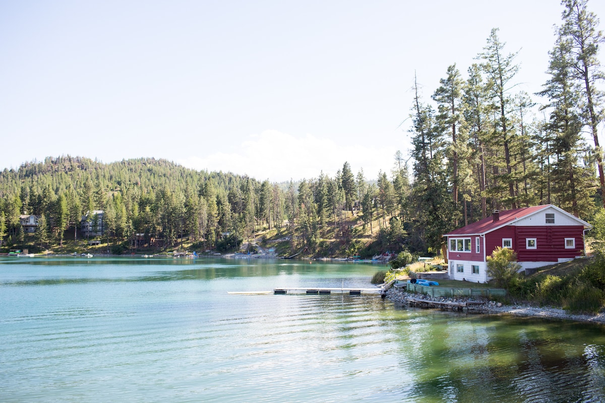 A charming red log cabin sits beside a tranquil lake, surrounded by lush green trees. The calm water reflects the natural landscape, with a private dock extending into the cove. The peaceful setting invites relaxation and exploration of the scenic surroundings.