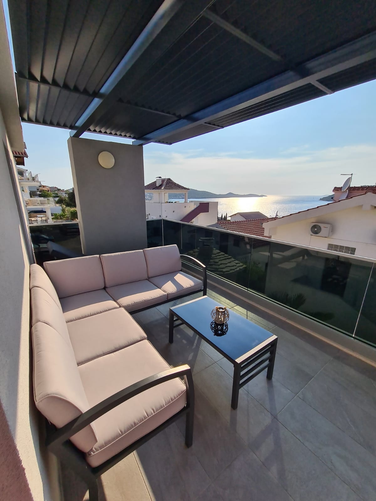 A shaded balcony is furnished with a light-colored seating arrangement and a small glass-top table. The view beyond extends to a serene sea, reflecting the sunlight. The surrounding buildings are visible, enhancing the sense of space and openness.