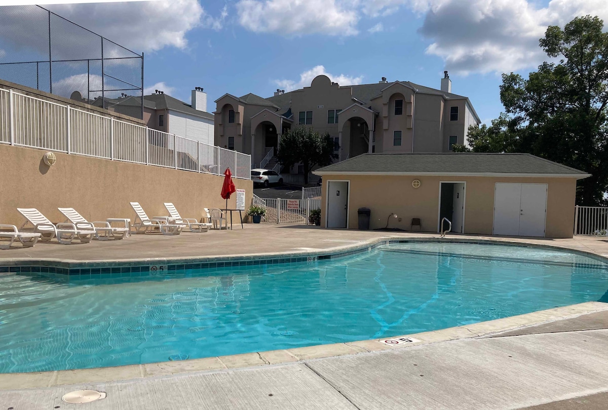 A clear swimming pool is visible, surrounded by a spacious patio with lounge chairs arranged neatly. A lifeguard station is situated nearby, and light clouds drift across a blue sky. In the background, the condominium building stands prominently.