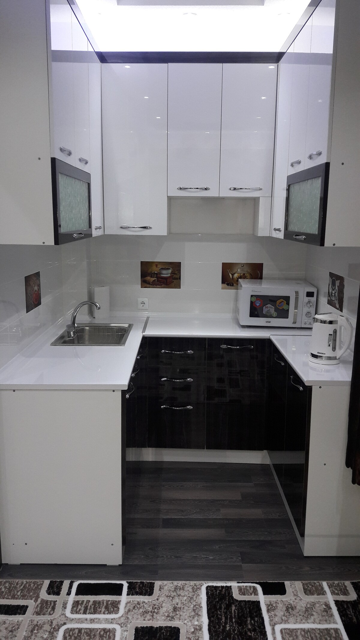 A compact kitchen features modern cabinetry in glossy white and dark wood. A stainless-steel sink is situated beneath a window. Essential appliances, including a microwave and kettle, are neatly arranged on the countertop, enhancing functionality within the space.