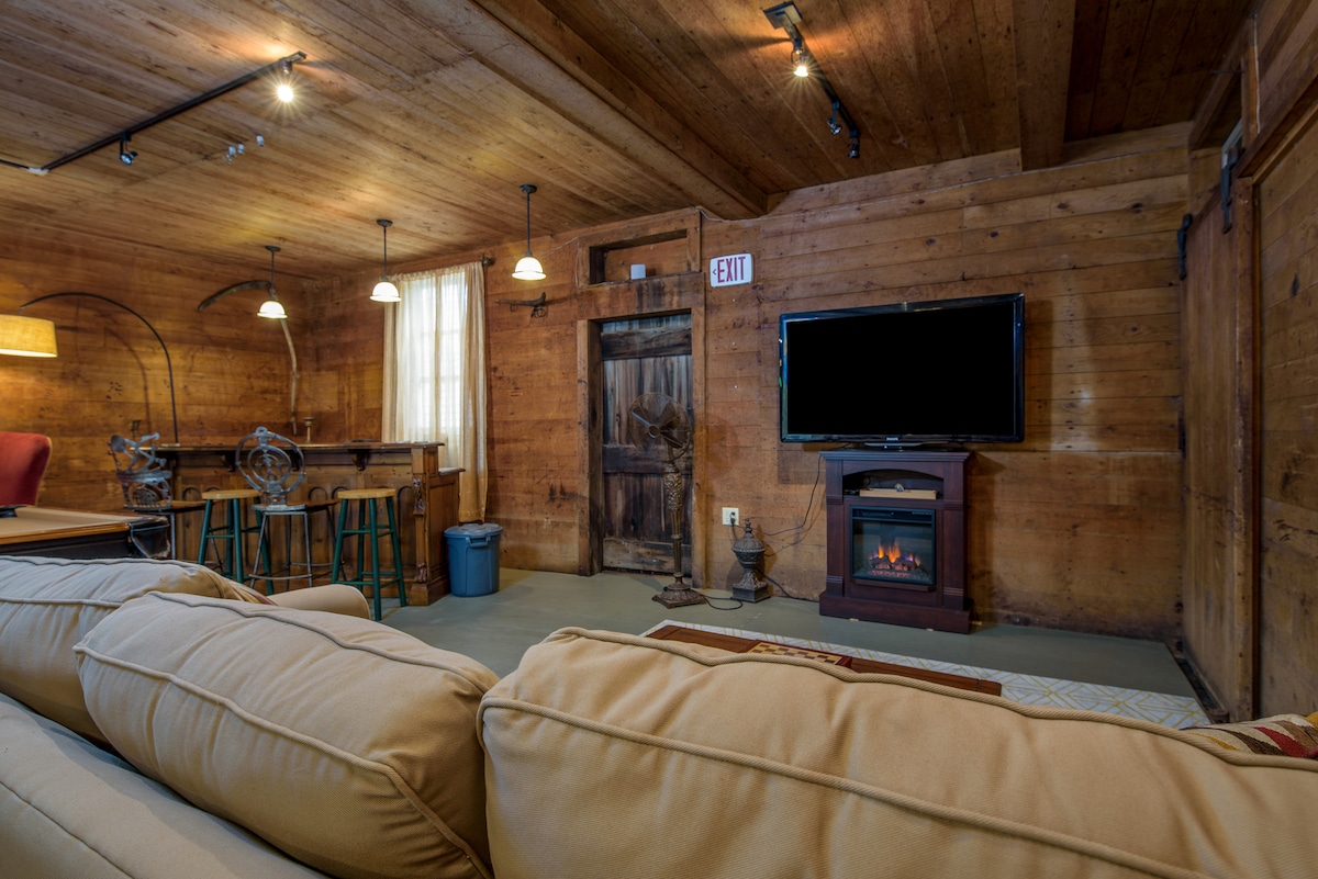 A cozy living area showcases wooden walls and ceiling beams, creating a warm ambiance. A large flat-screen TV is mounted on the wall, and a faux fireplace adds a touch of comfort. Bar-height stools are arranged near the exit, complementing the inviting seating area.