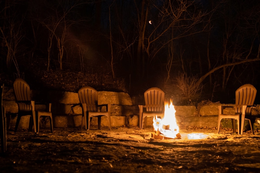 A cozy communal fire pit is surrounded by six wooden chairs, inviting relaxation under the night sky. The flickering flames illuminate the stone wall behind, creating a warm gathering spot amid the surrounding trees.