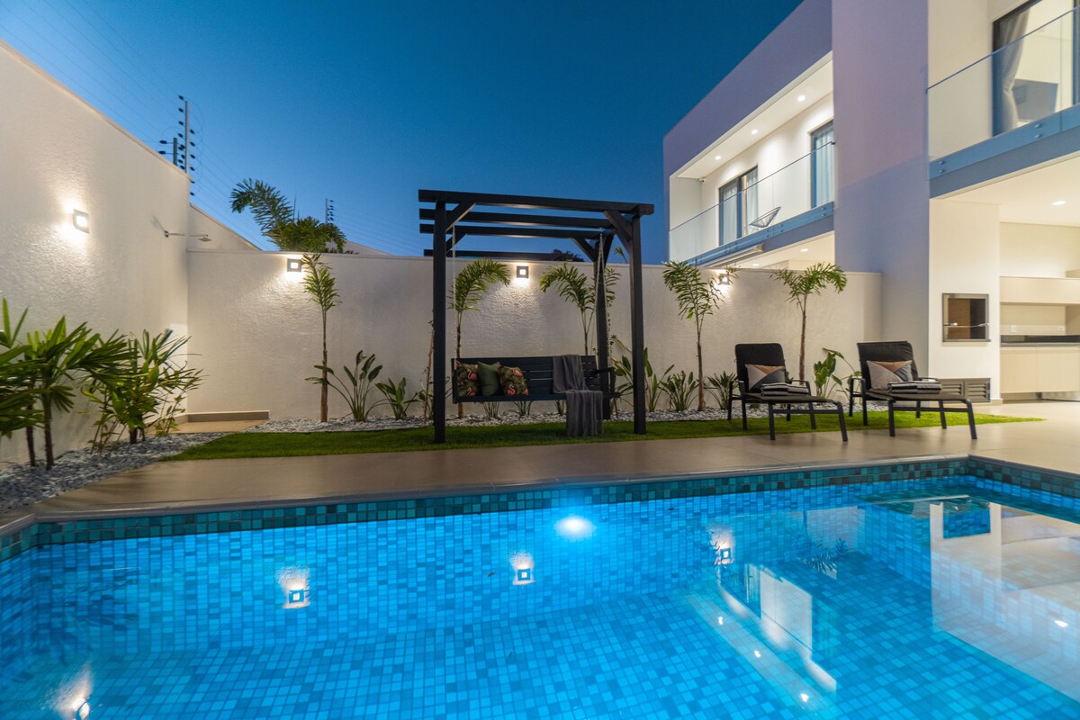 A private pool is surrounded by neatly manicured landscaping, featuring a wooden pergola with seating and lounge chairs nearby. The tranquil blue water reflects evening lights, while modern walls of the villa provide a clean and contemporary backdrop.