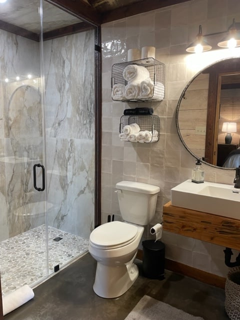 A modern bathroom features a walk-in shower with glass doors, showcasing elegant marble walls. A round mirror hangs above a wooden sink vanity. Stacked towels are neatly organized in wire baskets on the wall. A toilet is positioned adjacent to the shower.