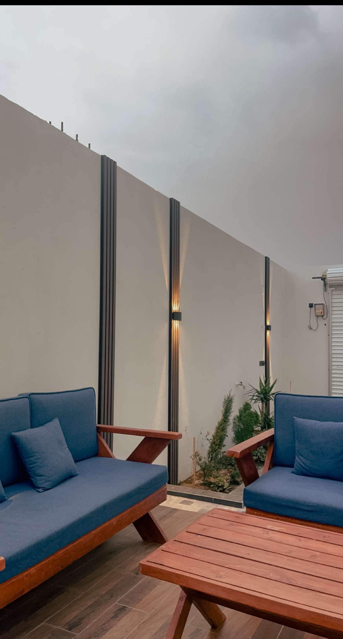 Two comfortable seating areas are arranged with blue cushions against a minimalist wall, where soft lighting enhances the ambiance. Small plants are positioned nearby, contributing to a relaxed outdoor environment.