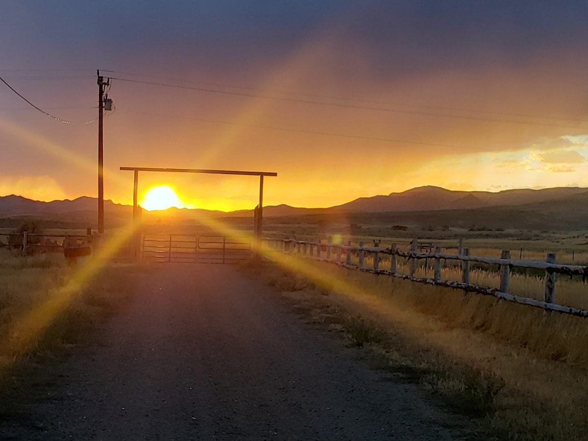 A serene landscape features a sun setting beyond a wooden gate, casting warm rays over a dirt road lined by a wooden fence. The horizon is filled with distant mountains, enveloped in soft hues of orange and yellow, creating a peaceful rural atmosphere.