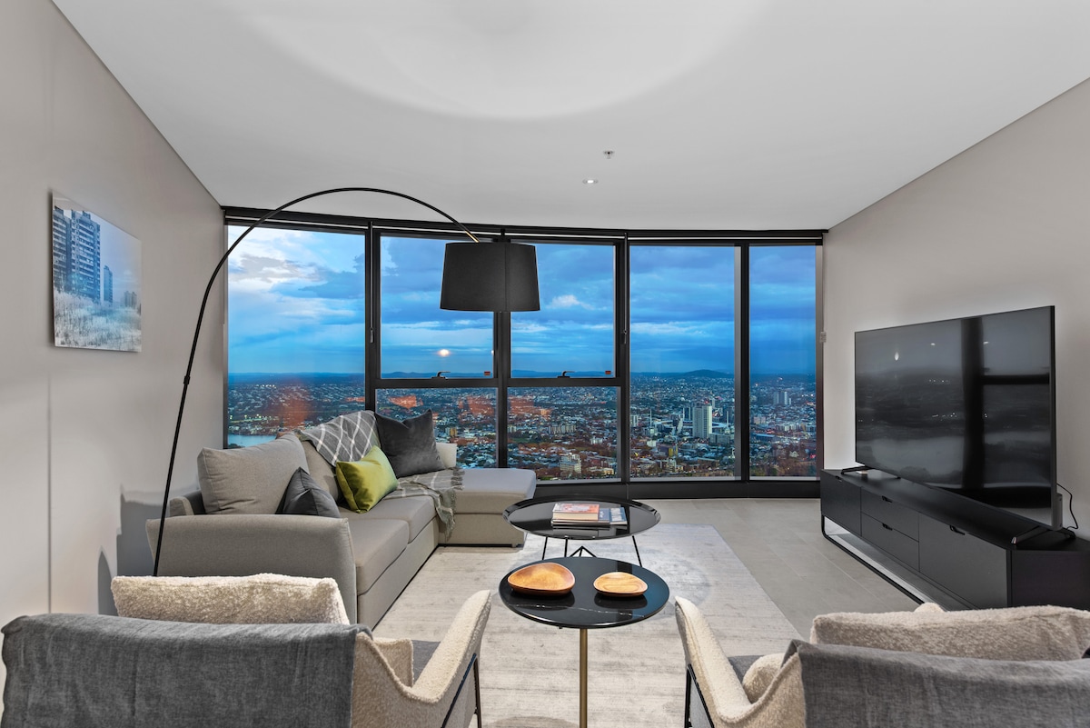 A modern living area features floor-to-ceiling windows offering sweeping views of Brisbane's skyline. A comfortable couch and two accent chairs surround a circular coffee table, with two plates placed atop. A large flat-screen TV is mounted on the wall, complementing the sophisticated decor.