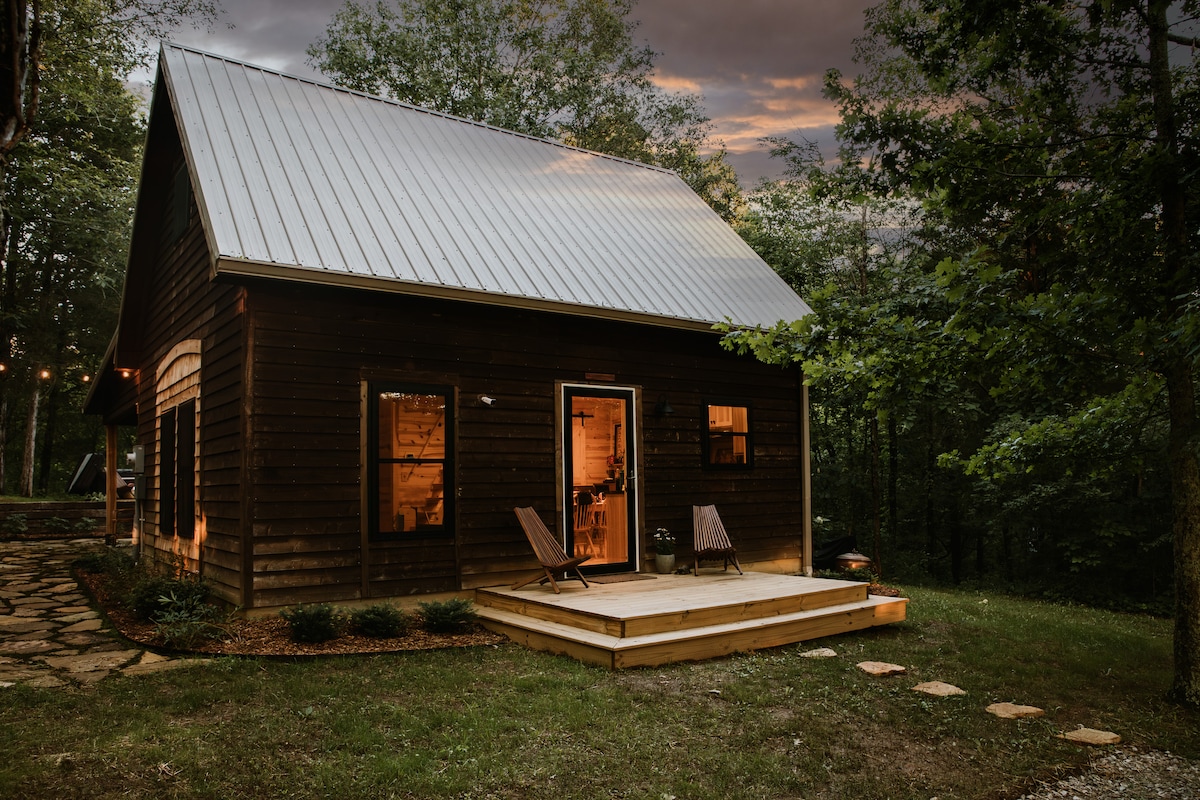 A rustic cabin is nestled among trees, featuring a well-lit entryway and a front deck with two wooden lounge chairs. Soft evening light spills through the windows, creating a welcoming atmosphere. Stone stepping stones lead towards the doorway, enhancing the natural setting.