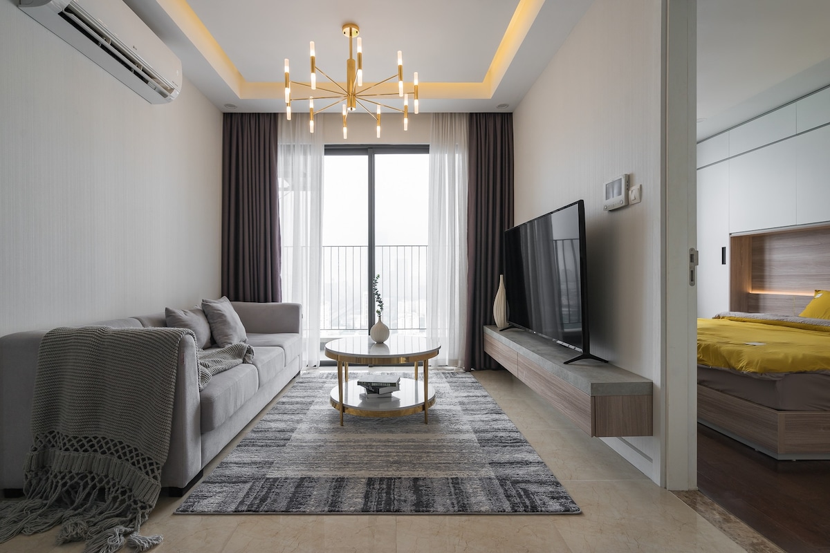 A modern living area features a light grey sofa complemented by a decorative throw. A round coffee table stands in the center on a patterned area rug. Natural light enters through large windows, while a mounted television is positioned against the wall.