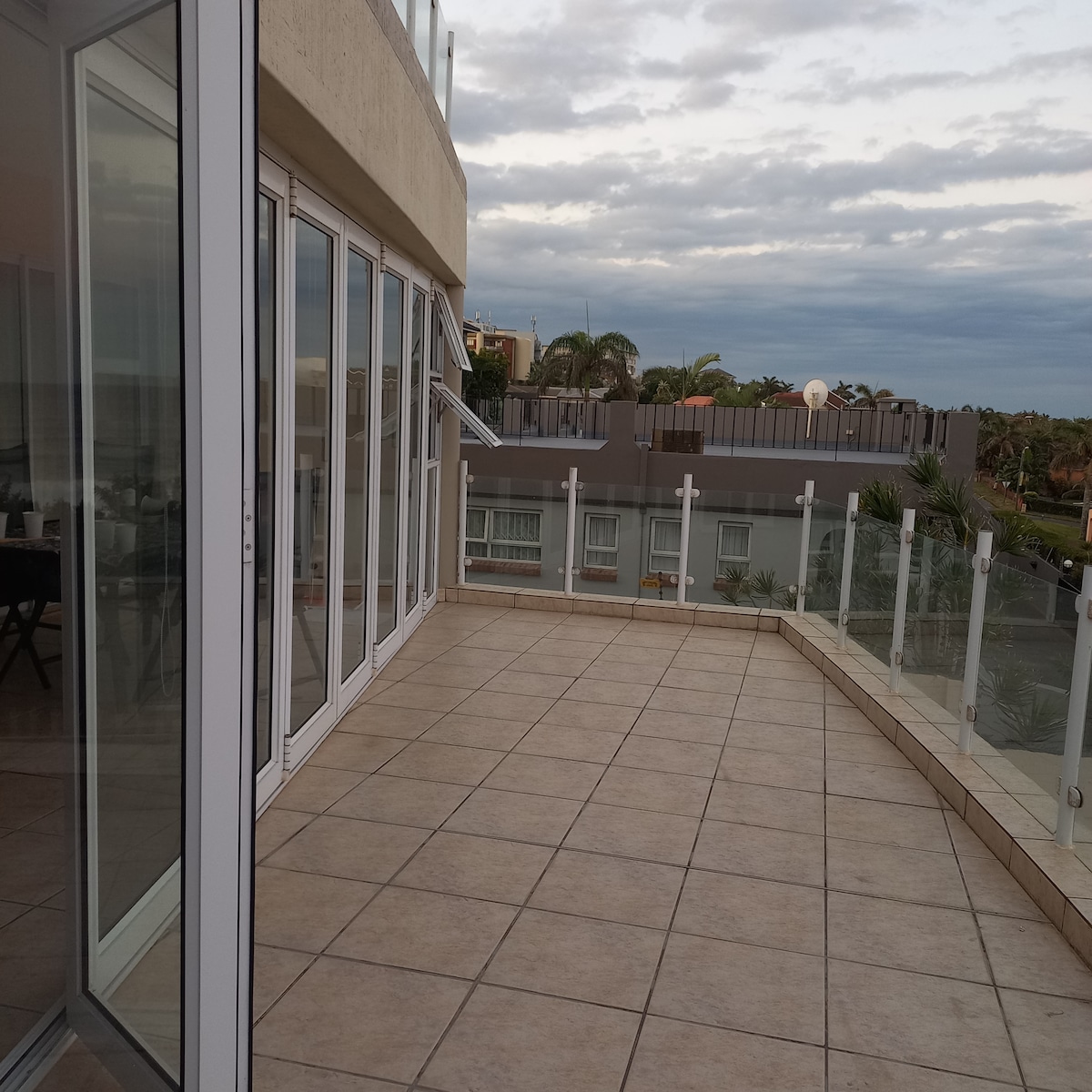 A spacious patio is framed by sliding glass doors, extending outdoor living space. Tile flooring is present, and a glass railing offers unobstructed views of the surroundings. Overcast skies are visible in the distance, creating a calm ambiance.
