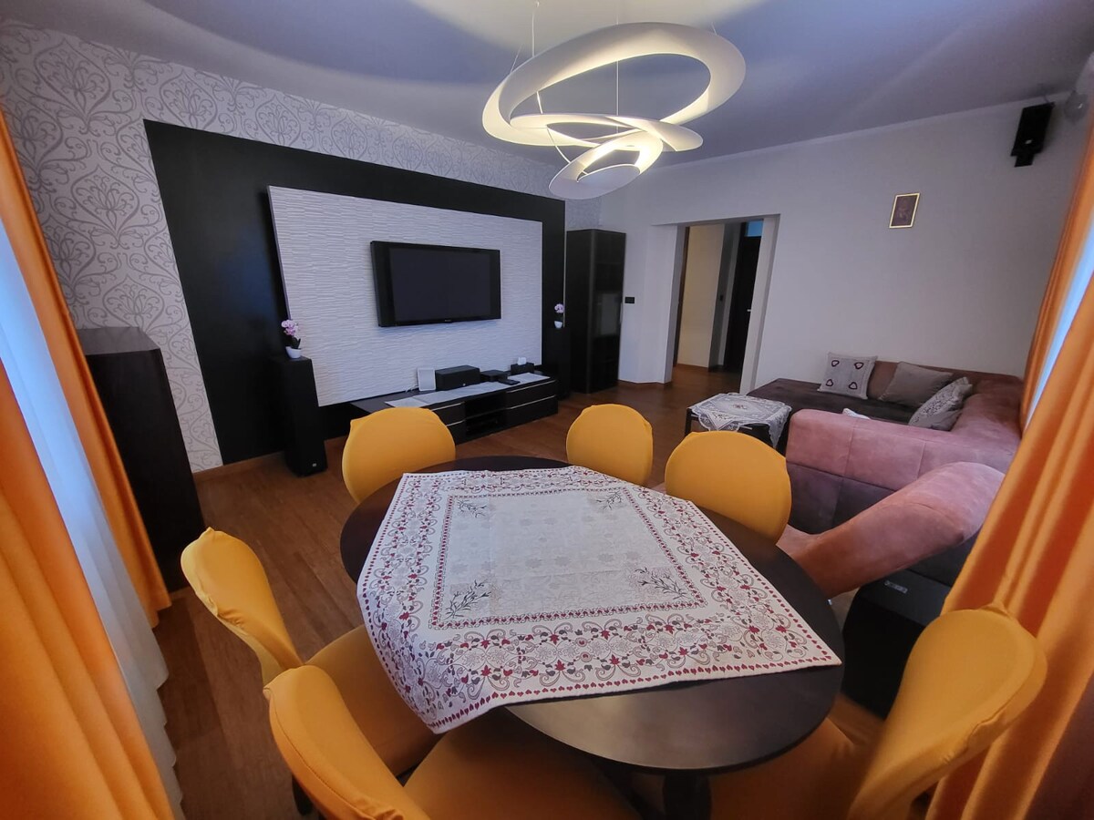 A spacious living area features a modern television mounted on a textured wall, complemented by a cozy couch. A round table surrounded by sunny yellow chairs is set at the center, adorned with a decorative tablecloth. Bright curtains frame the windows, allowing natural light into the space.