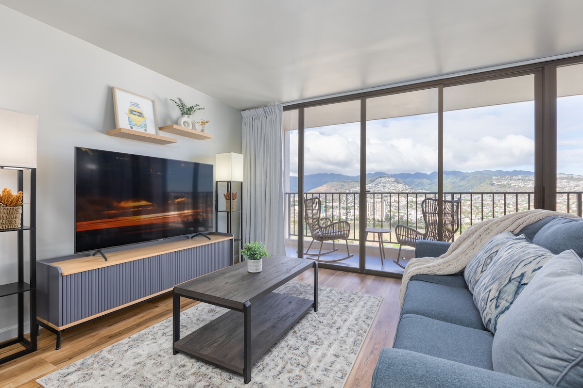 The living area features a spacious sofa in a calming blue hue and a wooden coffee table atop a patterned area rug. A large television is mounted on a wall shelf, with natural light streaming in from floor-to-ceiling glass doors that open to a scenic balcony with seating.