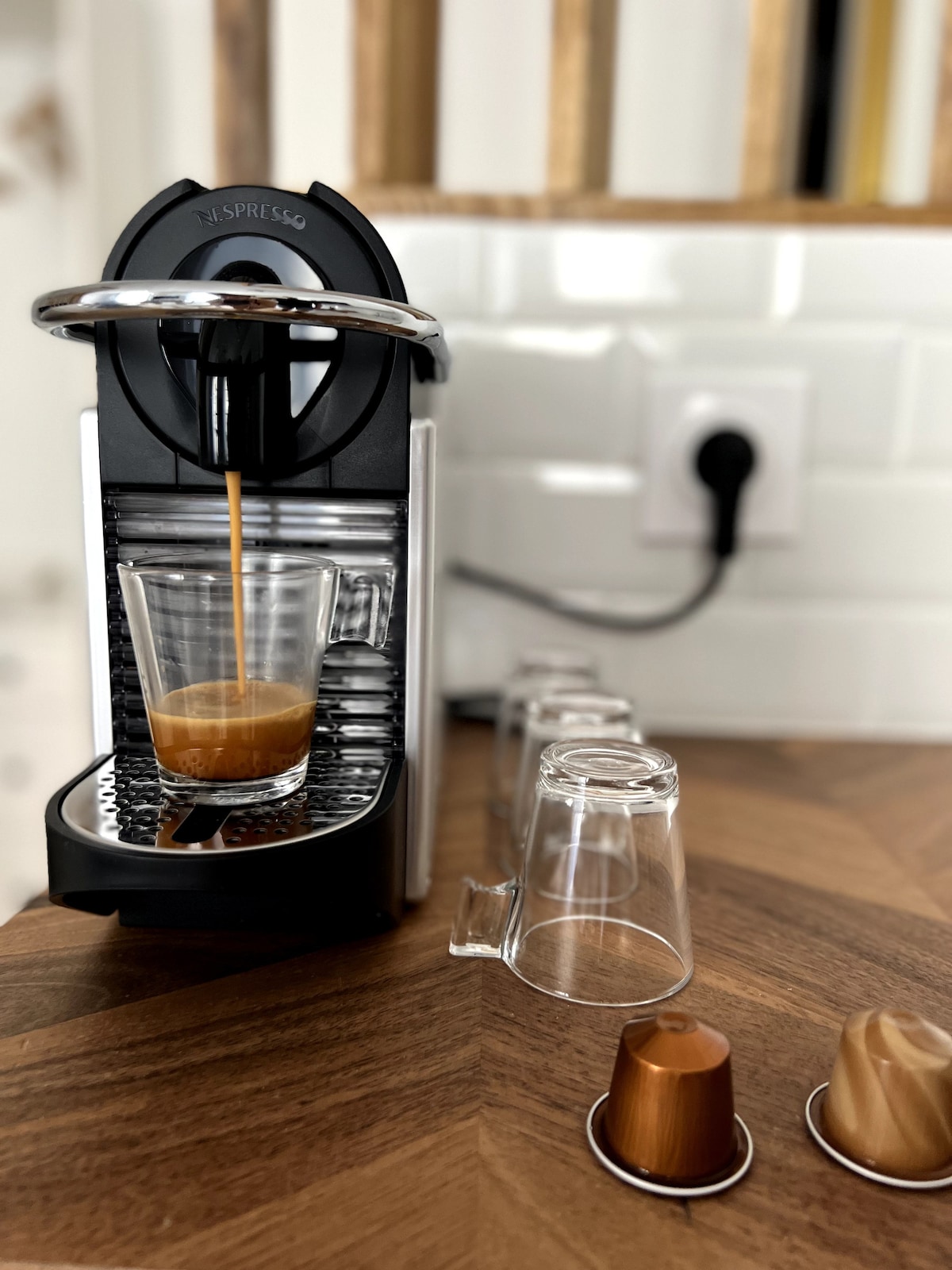 An espresso machine is seen dispensing coffee into a clear glass on a wooden surface. Several unused coffee capsules are displayed alongside small glass cups, with a power outlet in the background. The setup indicates a well-equipped kitchen area.