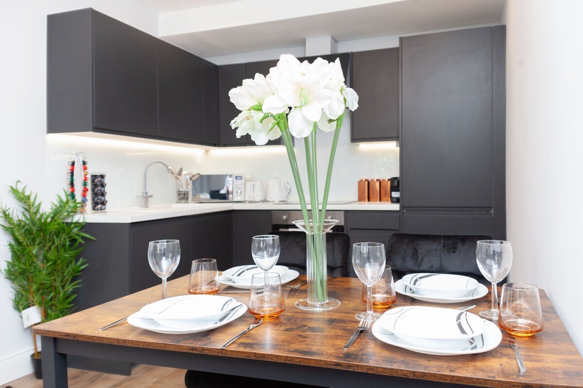 A modern kitchen is visible in the background, featuring sleek dark cabinetry and a white countertop. A wooden dining table is set for a meal, adorned with four plates, wine glasses, and glasses filled with amber liquid. A vase of white flowers adds a touch of elegance.