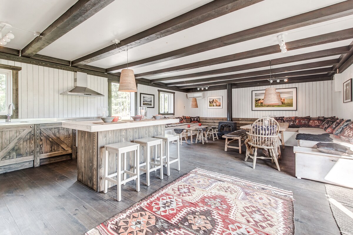 An open kitchen and lounge area is highlighted by wooden beams overhead. A large island with bar stools provides seating, while a cozy seating arrangement with cushions invites relaxation. Natural light enters through several windows, and a patterned rug adds warmth to the wooden floor.