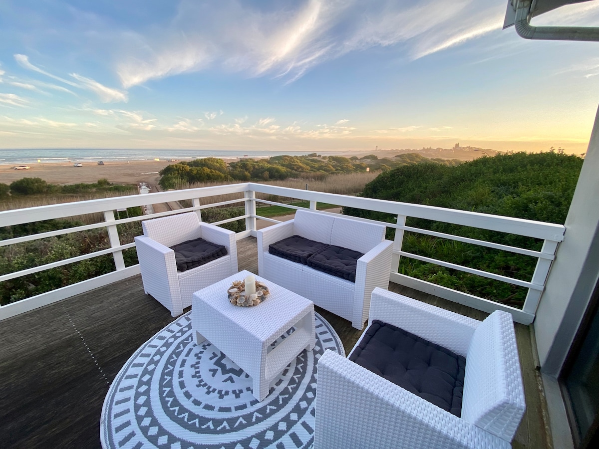 A seating area features four white wicker chairs contrasted with dark cushions, arranged around a small coffee table. The space is set on a deck overlooking a beach, with lush greenery and distant dunes visible under a colorful sunset sky.