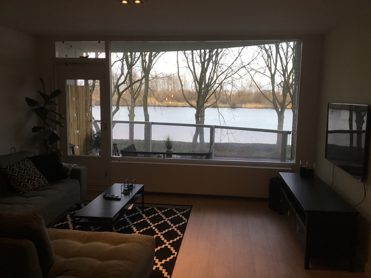 A bright living area features large windows that offer views of the water and surrounding trees. A comfortable couch is positioned beside a dark coffee table, and a television is mounted on the wall opposite. A patterned rug adds texture to the wooden flooring.
