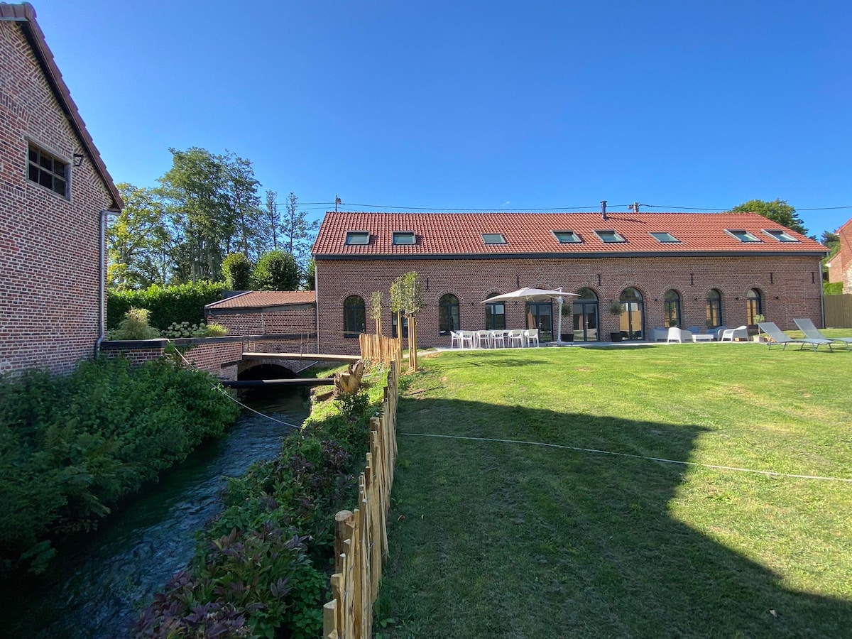 A spacious outdoor area is set alongside a flowing river, bordered by a wooden fence and lush greenery. A large building with multiple windows and a red roof stands in the background, complemented by a patio area with seating and a well-maintained lawn.