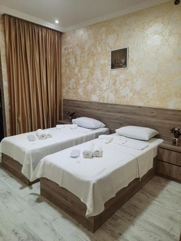 A well-lit guest room features two separate beds with crisp white linens and neatly arranged towels. A wooden nightstand sits beside each bed. Decorative wall art is displayed above the headboard, while soft drapes frame a window, allowing natural light to brighten the space.