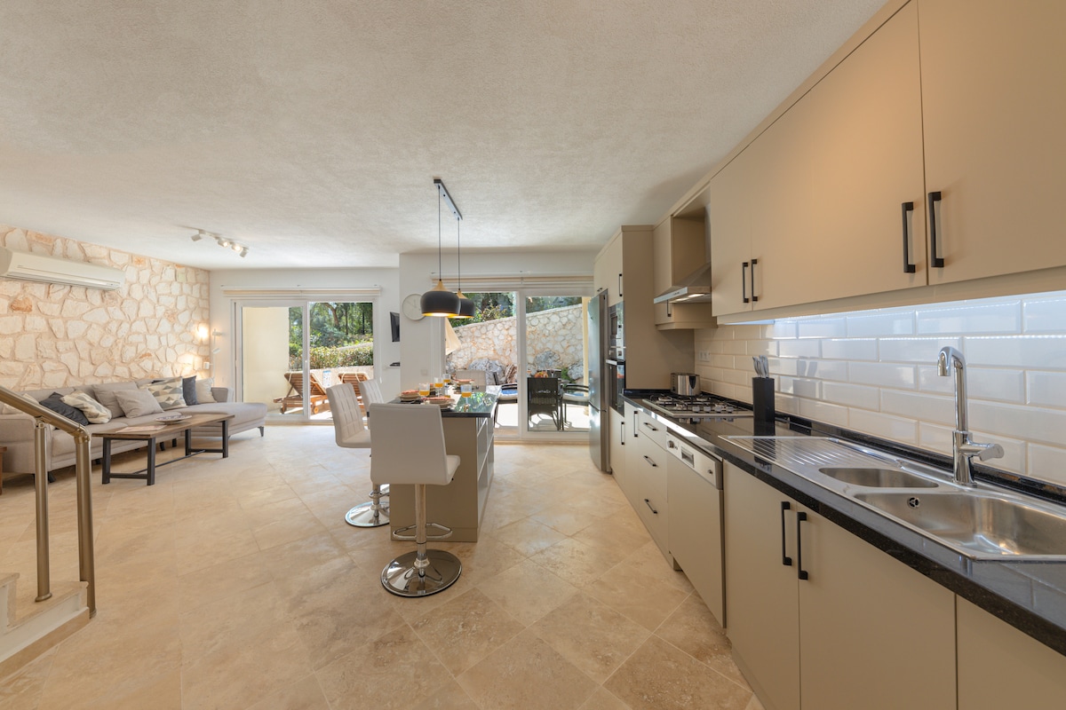 An open-plan living area is shown, featuring a modern kitchen with sleek cabinetry and stainless-steel appliances. A large island with stools sits at the center, while a dining area with a table and chairs is visible. Natural light enters through patio doors leading to an outdoor space.
