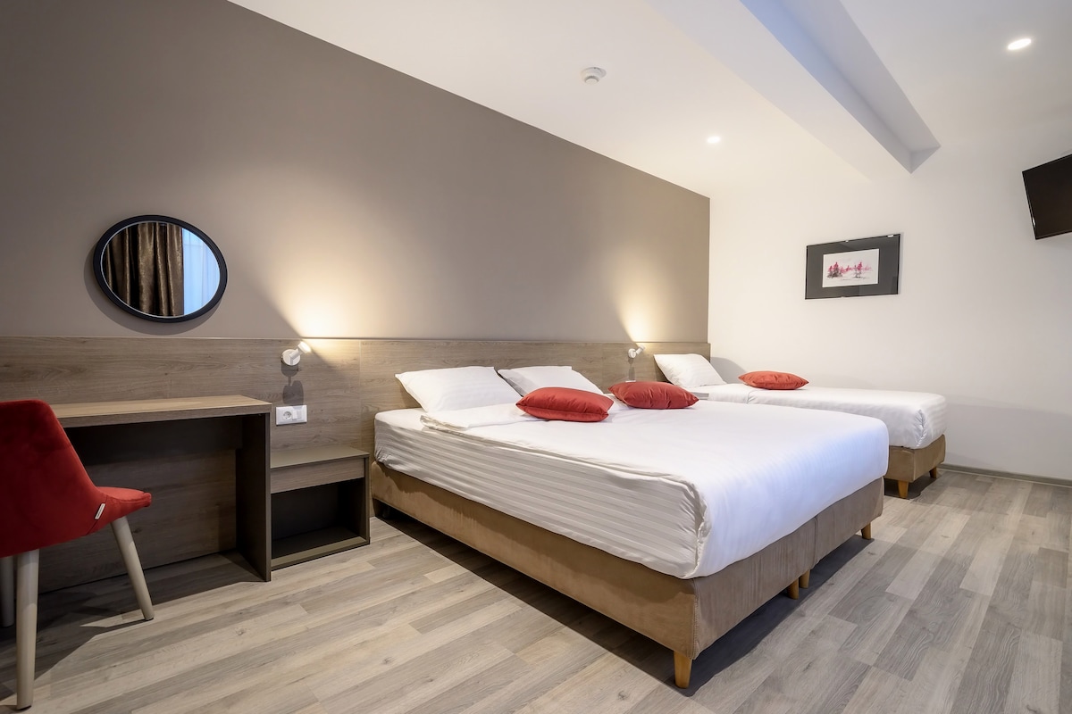 A modern room is furnished with two spacious beds and decorative red accent pillows. A small desk with a chair is positioned against the wall, complemented by a circular mirror. Soft lighting highlights the wooden flooring and neutral wall tones.