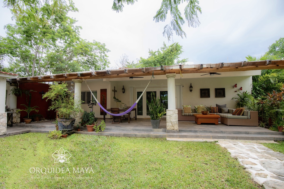 A welcoming terrace is displayed with comfortable seating arrangements and hammocks. Lush greenery surrounds the area, enhancing the serene atmosphere. Planters filled with various plants create a vibrant setting while the roof provides shade. A stone path leads into the inviting space.