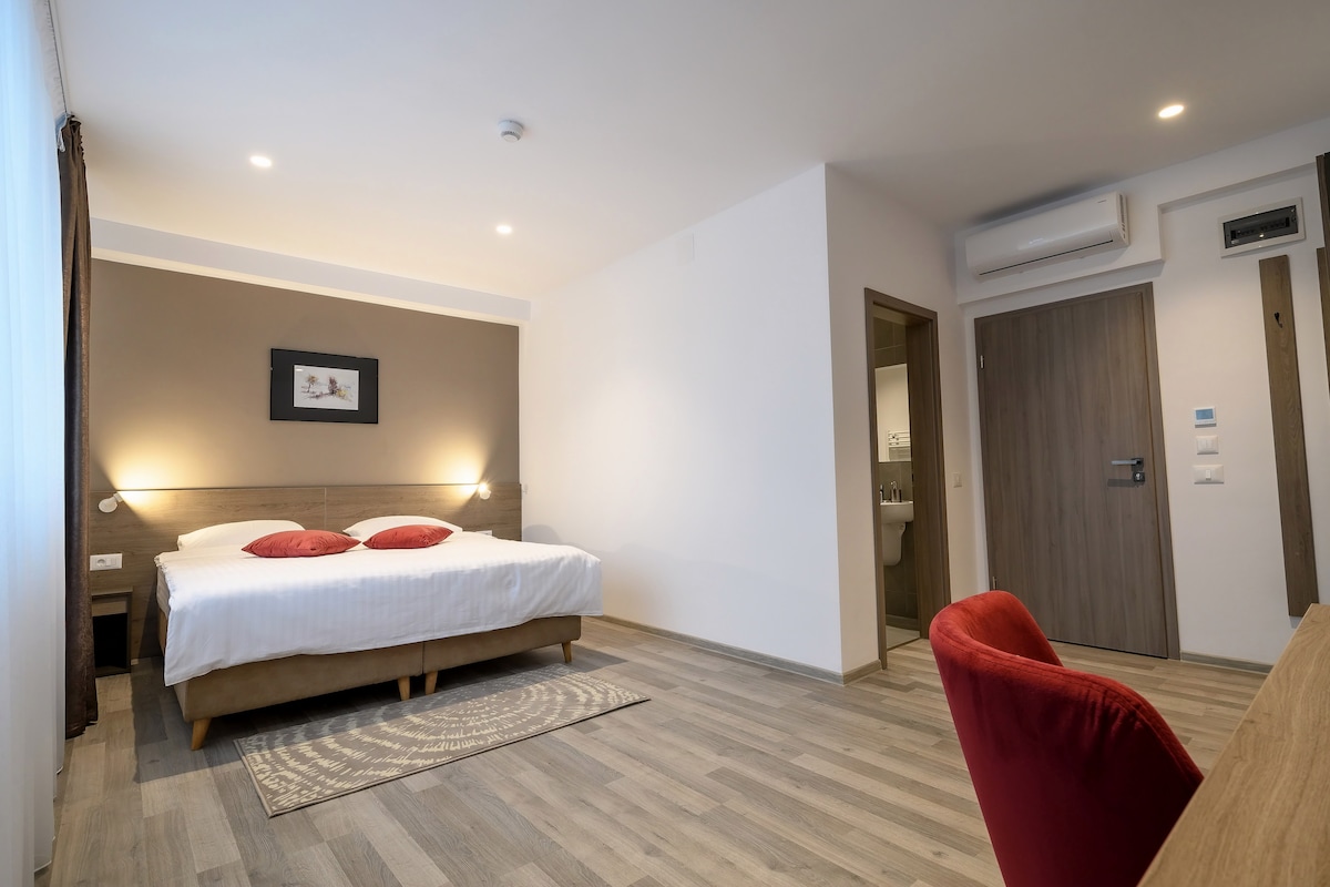 A modern guest room is presented with a spacious bed adorned with red accent pillows. A simple wooden desk and a red chair are placed nearby. Natural light enters through a large window, while soft lighting enhances the neutral-toned walls and wooden flooring.