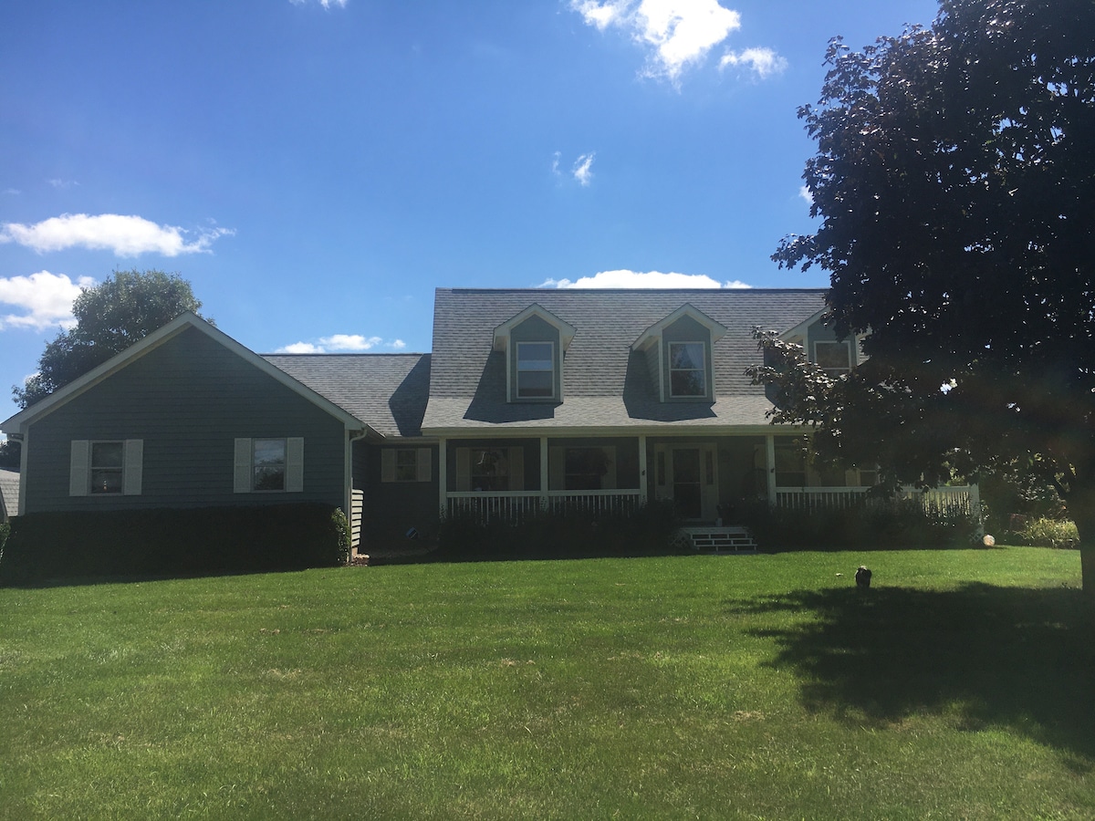 A Cape Cod style home is featured with a sloped roof and multiple gabled windows. A large front lawn extends towards the house, which is surrounded by greenery. The facade is painted in a soothing green hue, complemented by white trim and a welcoming porch.