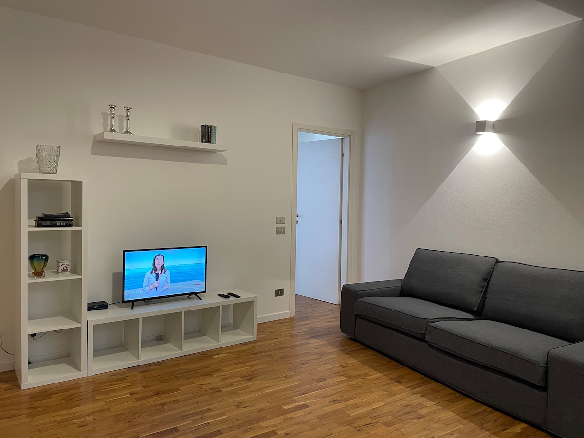 A living area is depicted featuring a gray sofa and a television placed on a white shelving unit. Natural wood flooring adds warmth, while a decorative vase and additional items on the shelves create an organized space. Soft lighting enhances the room's brightness.