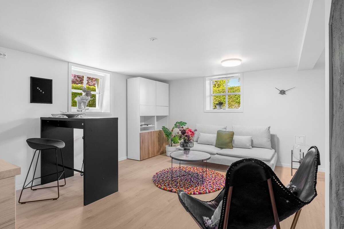 A bright and airy living area features light walls and wooden flooring. A cozy sofa is positioned beside a glass coffee table on a colorful circular rug. A small dining table with high chairs is present, while two windows provide natural light and views of greenery.