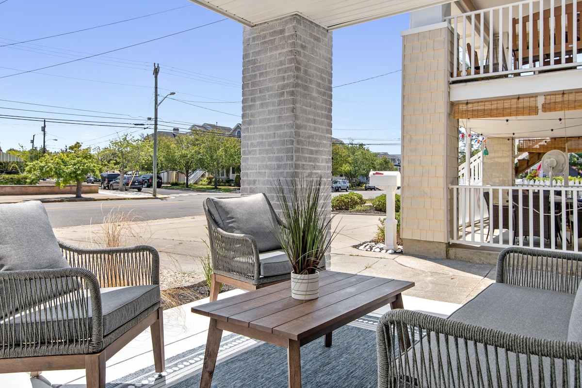 An outdoor patio seating area is showcased, featuring two modern armchairs and a low coffee table. Potted plants add a touch of greenery. Views of the neighborhood can be seen in the background, highlighting the accessibility of nearby attractions.