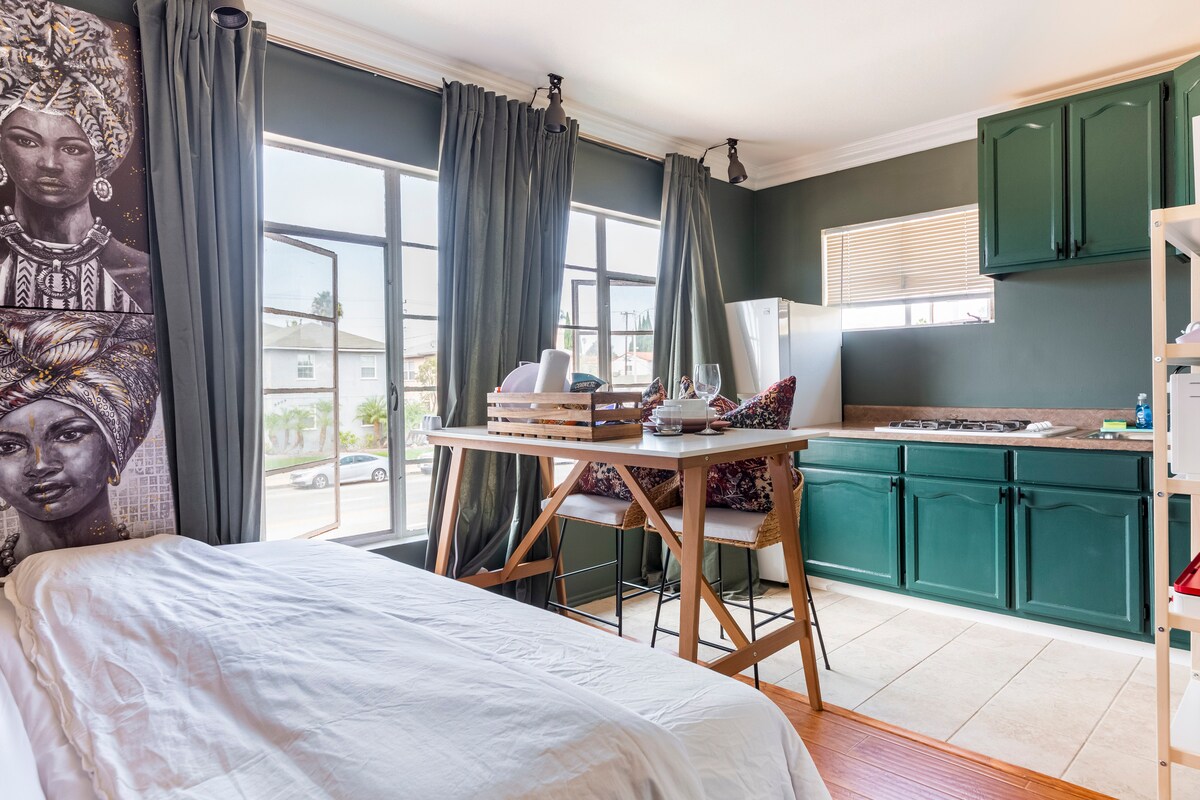 Natural light fills the cozy studio, highlighting the spacious bed with crisp linens. A small dining area is set with dishes and utensils, adjacent to a kitchenette featuring green cabinets and a refrigerator. The large windows are framed by dark curtains, enhancing the contemporary decor.