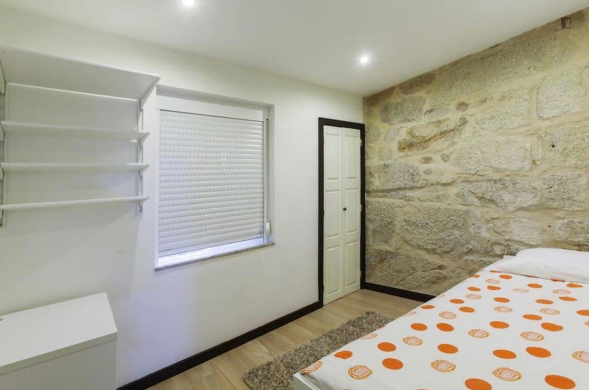 A cozy room features a double bed with an orange polka dot patterned blanket. Stone walls add character to the space. A small shelf and a simple white cupboard are positioned against the wall, while a window with blinds allows natural light to enter.