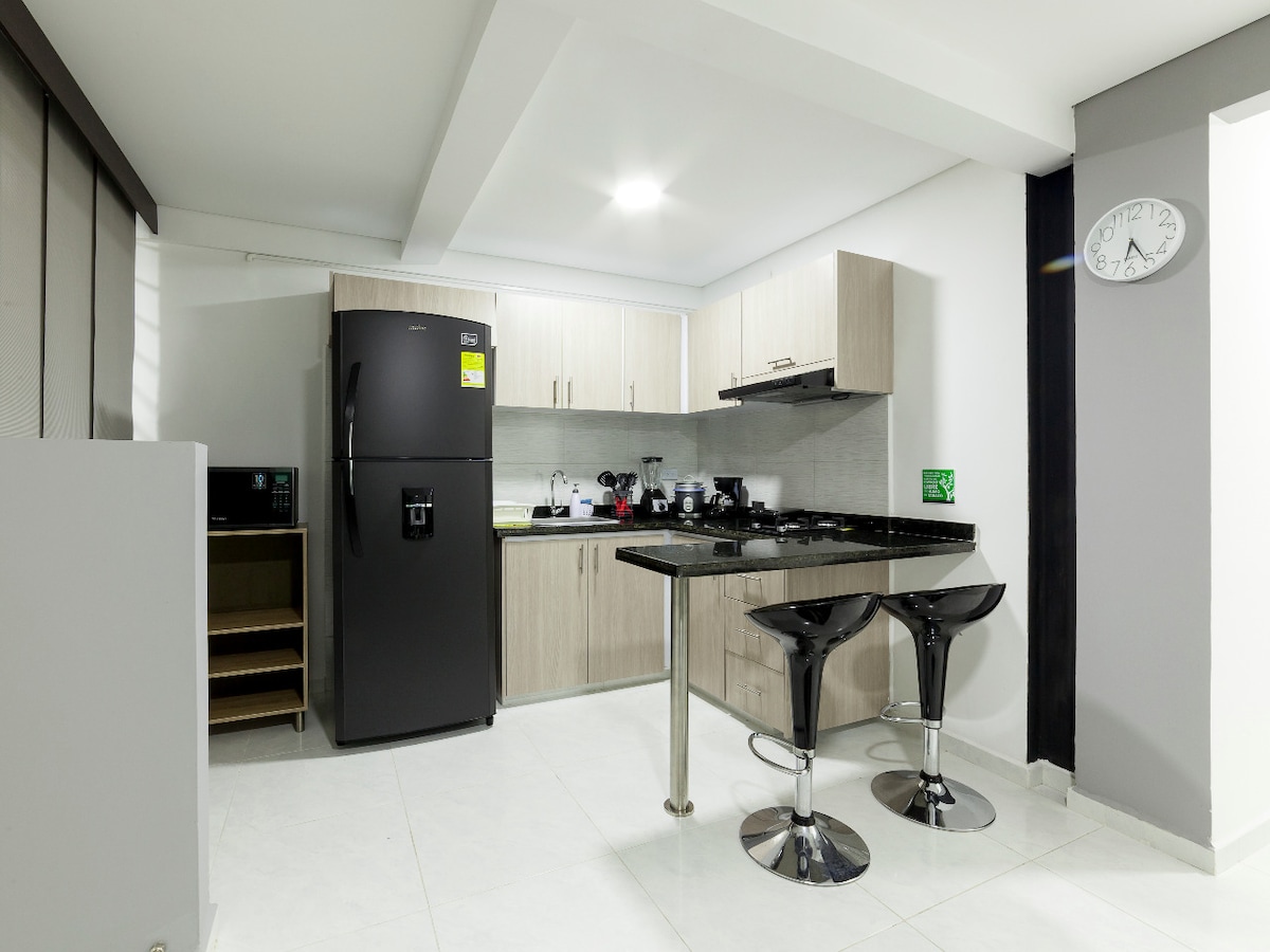 A modern kitchen is displayed, featuring sleek cabinetry and a black countertop. Appliances include a refrigerator, microwave, and stove, along with various kitchen utensils. Two stylish bar stools are positioned at a small dining table, complementing the contemporary design.