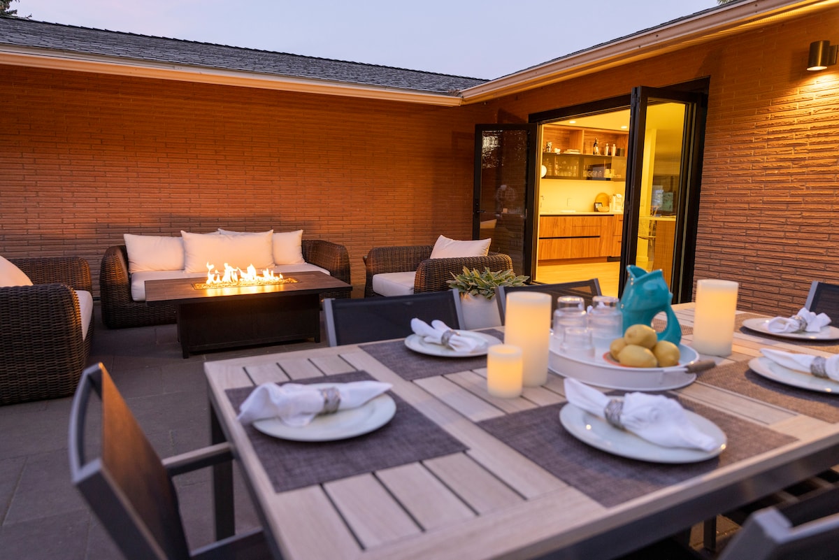 An outdoor dining area is featured with a wooden table set for six, including white plates and linen napkins. A gas fire table provides warm illumination, surrounded by comfortable seating in neutral tones. Glass doors lead to a modern kitchen, enhancing the inviting atmosphere.