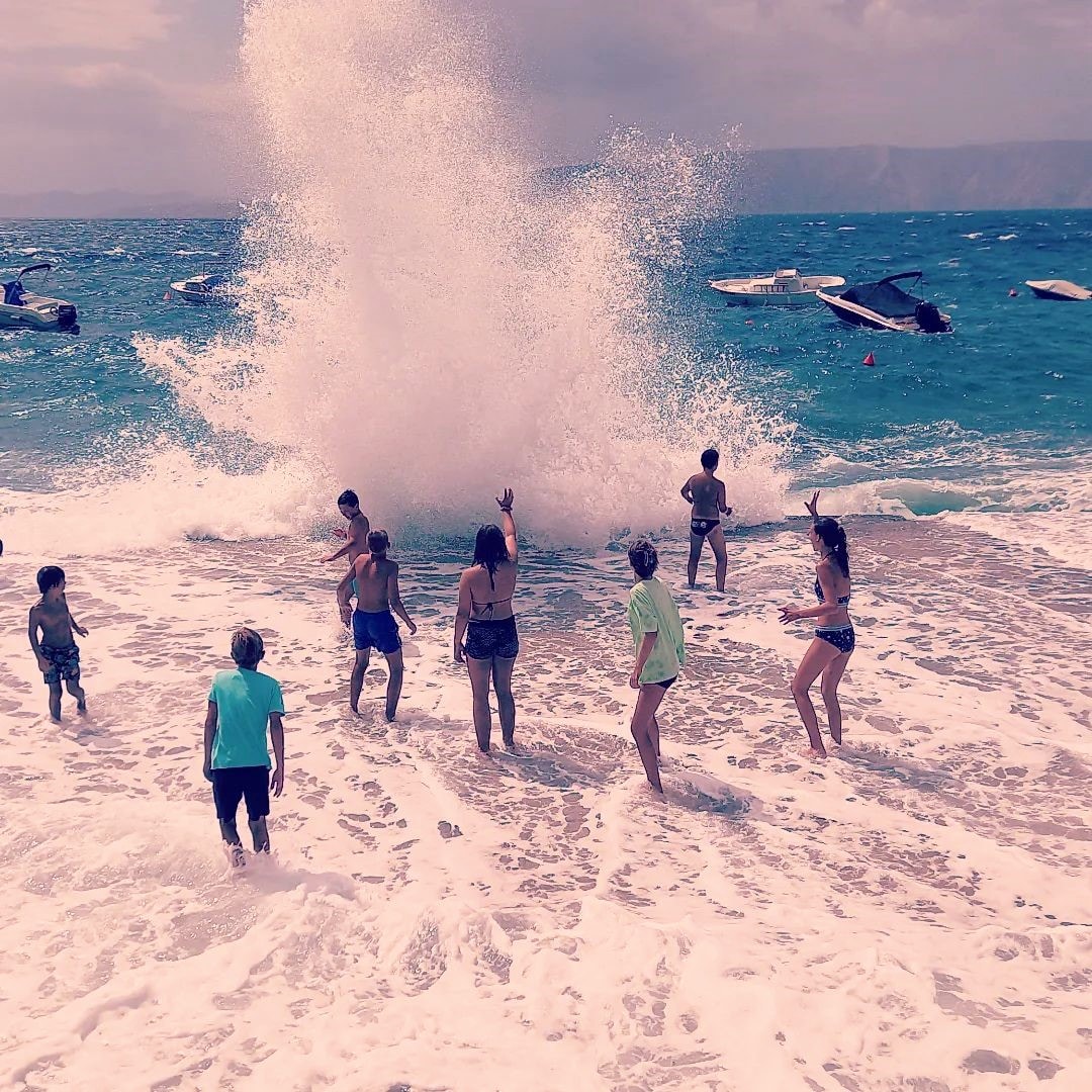A lively beach scene is captured as waves crash onto the shore, creating a splash. Several people, including children, are seen interacting with the water, wearing swimwear. Nearby boats are visible in the crystal blue sea, adding to the beach atmosphere.