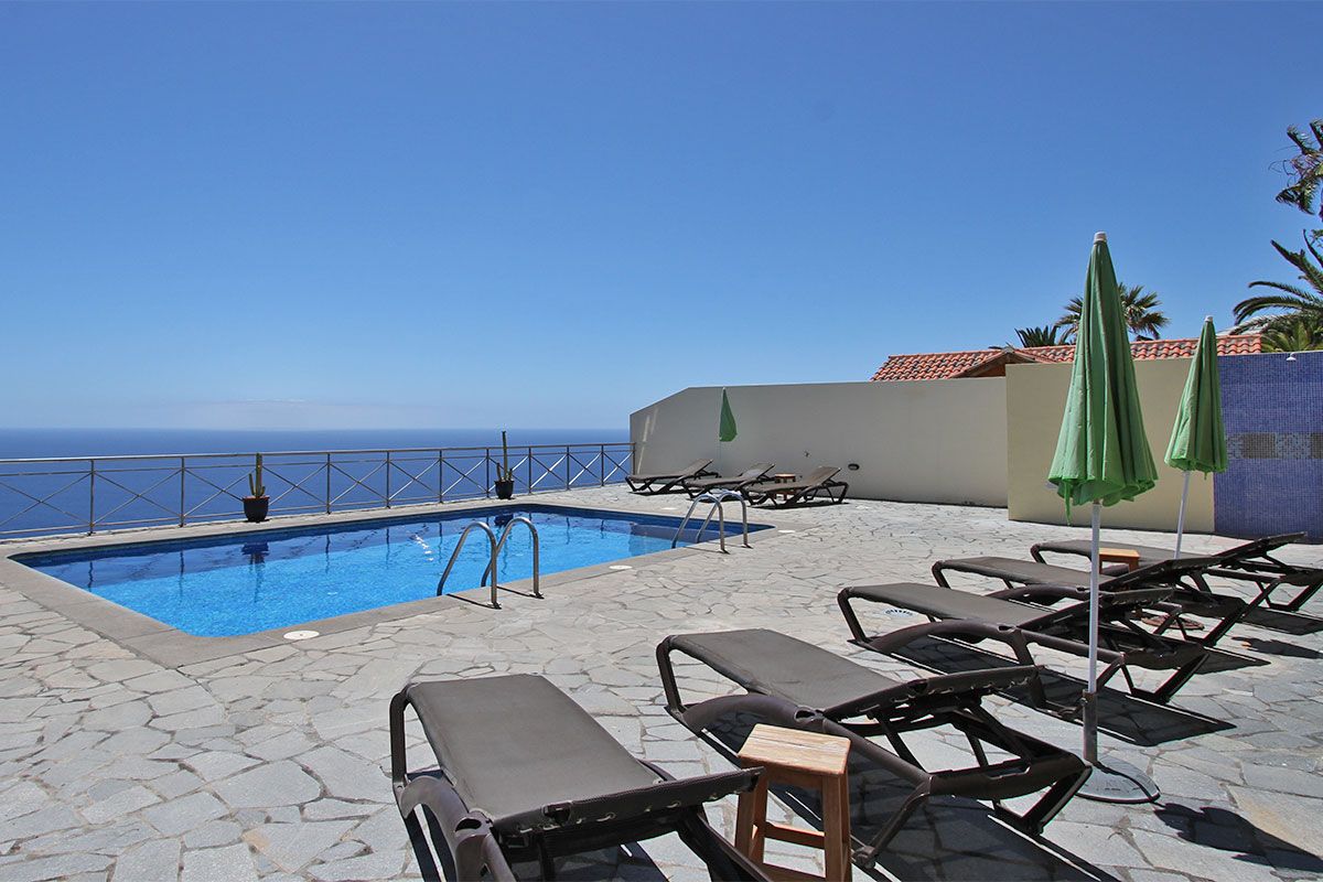 An outdoor area features a rectangular swimming pool surrounded by a stone deck. Several lounge chairs are arranged around the pool, complemented by two green umbrellas. A distant view of the ocean is visible beyond a low wall.