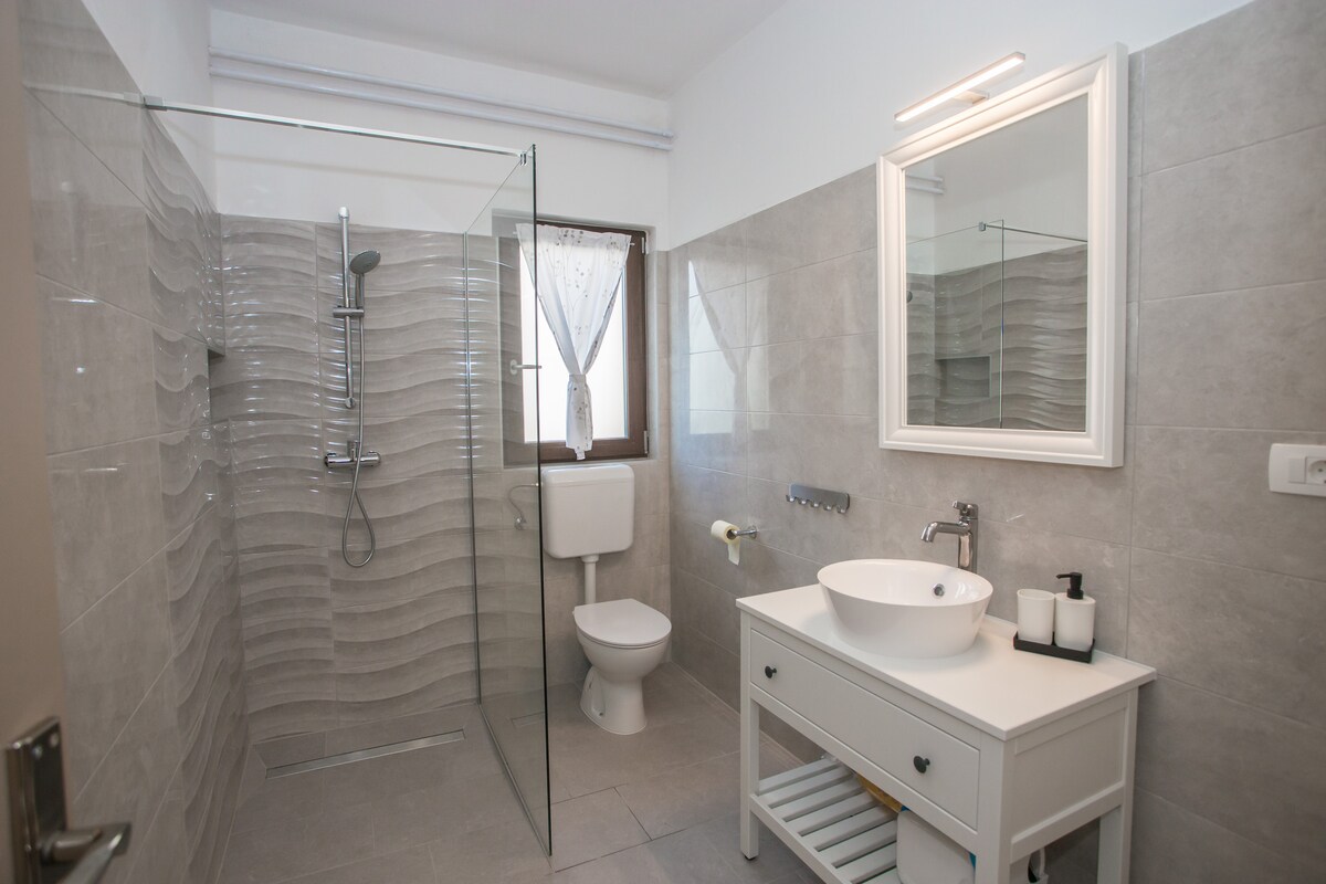The bathroom features modern fixtures with a walk-in shower, equipped with a rain showerhead. A sleek white vanity with storage space showcases a circular sink. Natural light enters through a window, which is adorned with sheer curtains. Neutral tiles create a clean and spacious feel.