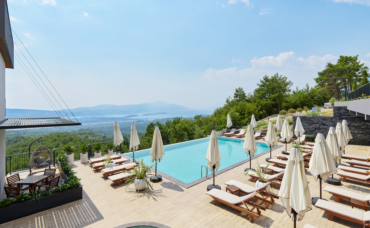 An outdoor pool area is surrounded by sun loungers beneath large umbrellas. Lush greenery and distant hills are visible in the background, complemented by a clear blue sky. The pool reflects the sunlight, creating a serene atmosphere for relaxation.