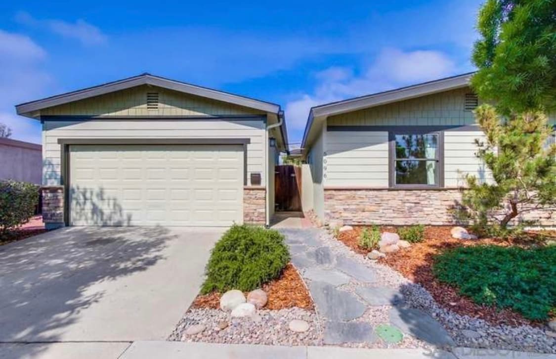A modern, single-story residence features a two-car garage with a smooth driveway leading to the entrance. Landscaping is showcased with bushes and stone paths, creating a welcoming approach. Blue skies and soft clouds provide a serene backdrop to the well-maintained exterior.