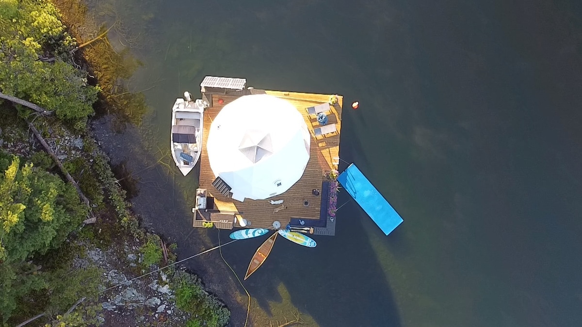 An aerial view showcases a floating yurt surrounded by water. The circular white yurt is complemented by a wooden deck, featuring two paddle boards and a canoe resting on the edge. Lush greenery frames the scene, enhancing the secluded natural setting.