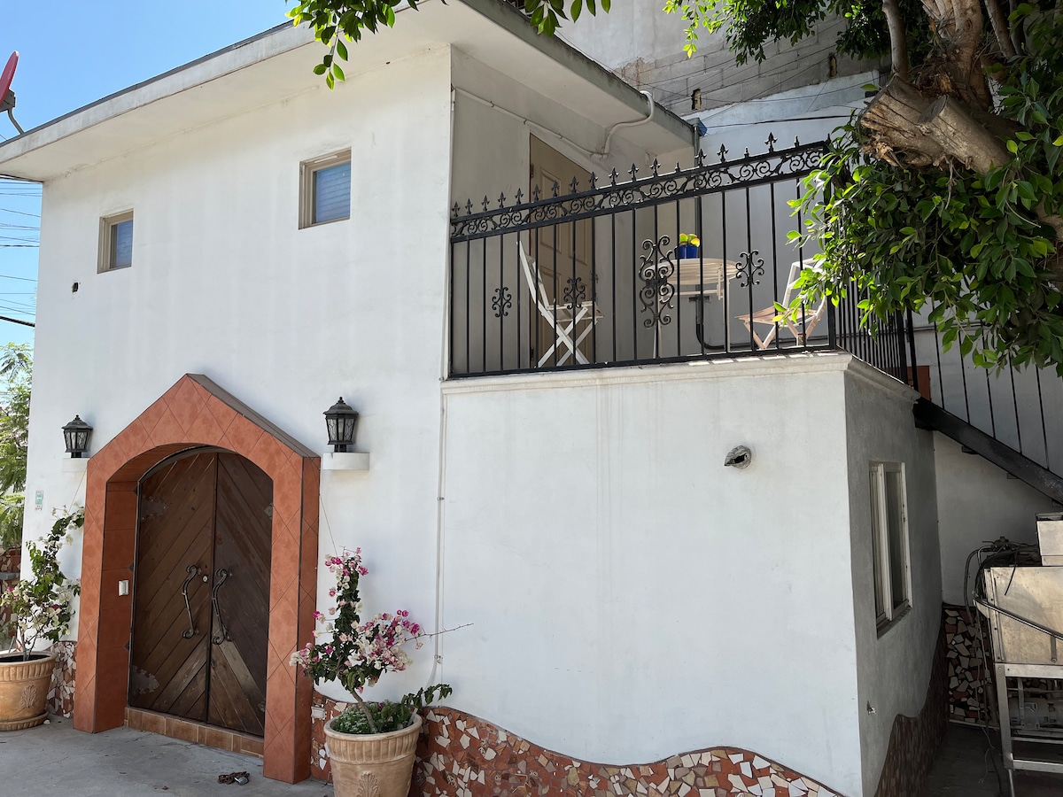 A two-story structure is presented with a welcoming entrance featuring a large wooden door. A small balcony extends from the upper level, adorned with white chairs and surrounded by lush greenery and potted plants.
