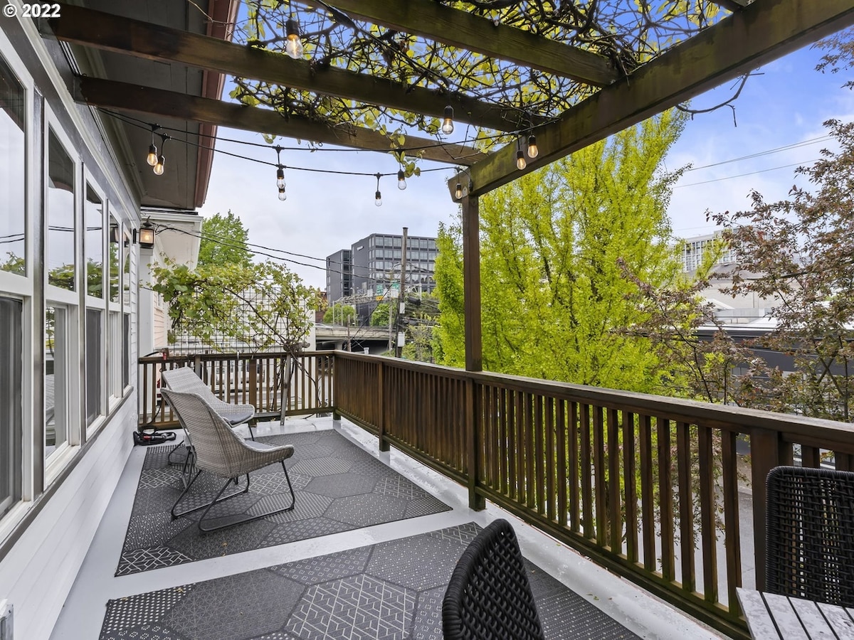 An outdoor deck area features a wooden railing and hanging string lights. Two lounge chairs are positioned for relaxation, while a dining table with chairs is set up for gatherings. Verdant greenery and a nearby building are visible in the background, enhancing the inviting outdoor space.