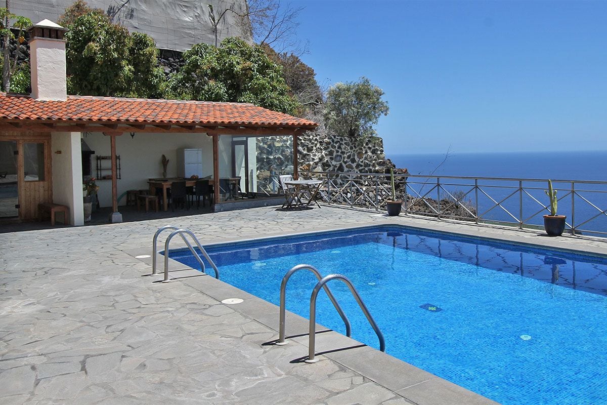 A serene outdoor space features a sparkling blue pool, surrounded by a stone patio. A covered area with wooden beams and a tile roof includes a dining table and chairs. Lush greenery enhances the backdrop, providing a scenic view of the ocean.