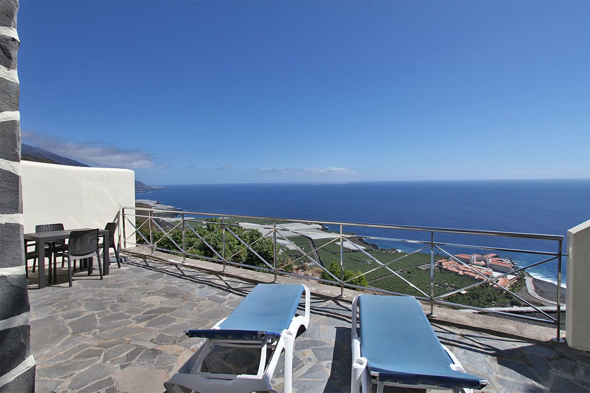 A spacious terrace features two loungers oriented towards an expansive ocean view. A four-person dining table stands nearby, positioned against a backdrop of coastal scenery. Clear blue skies complement the tranquil atmosphere, and vibrant greenery is visible in the landscape below.