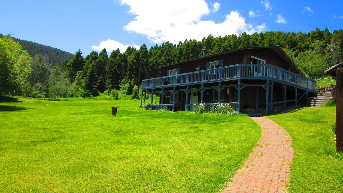 A spacious house is shown surrounded by vibrant green grass and towering trees. A brick pathway leads to the entry, and a large deck wraps around the home, providing ample outdoor space for relaxation. The clear blue sky adds to the serene environment.